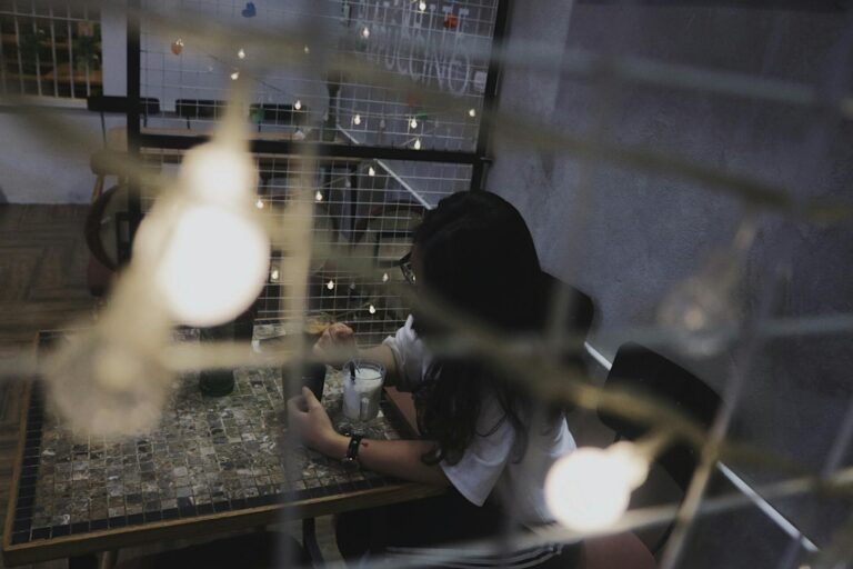 A woman sits alone at a table in a dimly lit café with soft, blurred lights creating a cozy atmosphere.